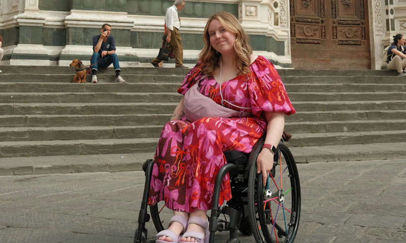 Jasmin, is in a wheelchair smiling in front of a historic building. She has long blonde hair, is wearing a hot pink dress, and pink platform sandals.
