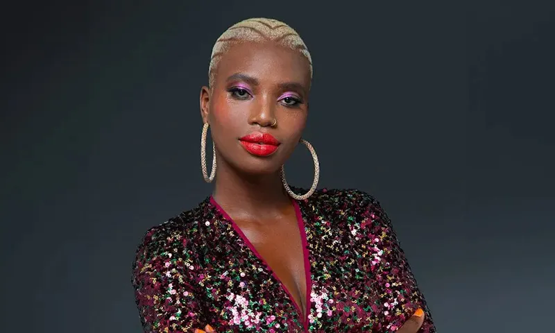 Khadija, a disability advocate, stands with arms folded. Khadija has short blonde hair and is dressed in a sequence top.