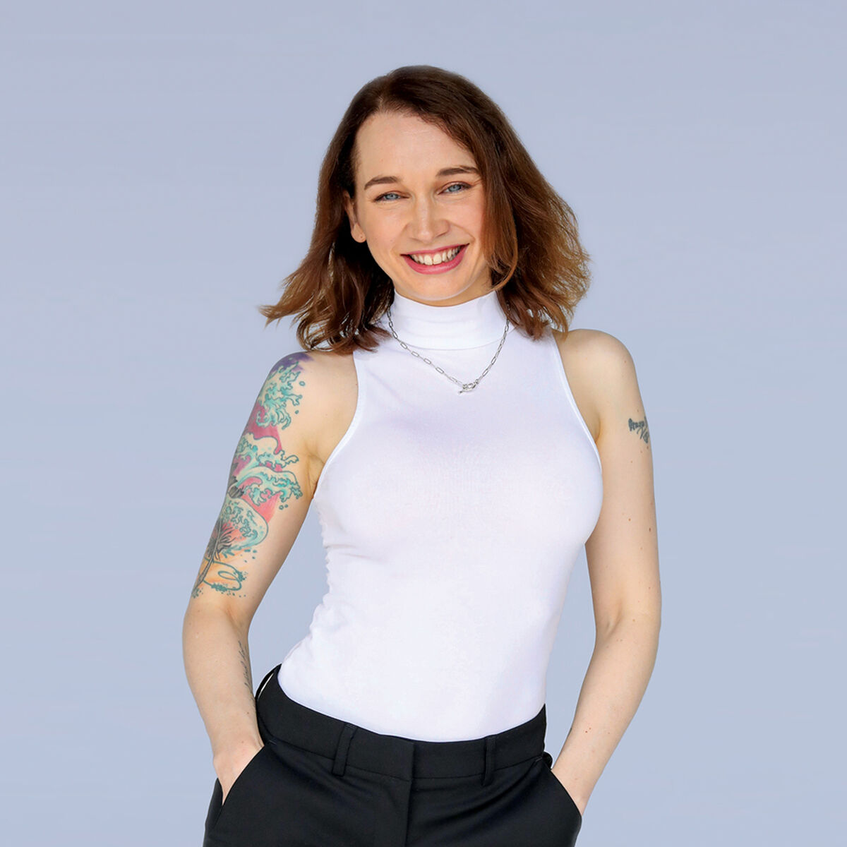 Flic is standing in front of a white backdrop. She wears a white sleeveless tunic and has a tattoo on her left shoulder. 