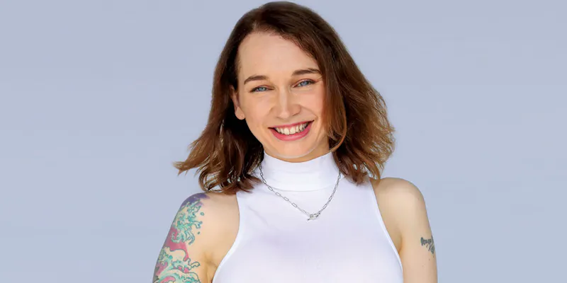 Flic is standing in front of a white backdrop. She wears a white sleeveless tunic and has a tattoo on her left shoulder.