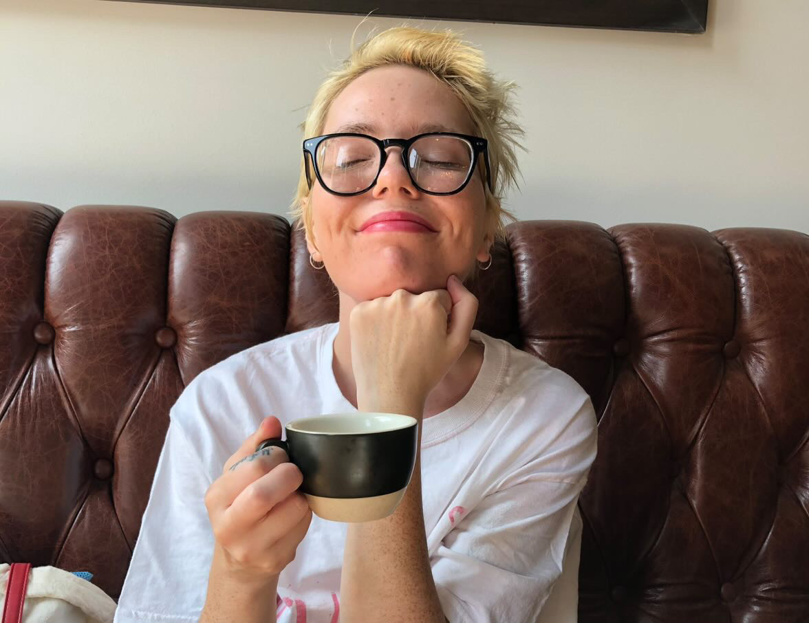 Amber is sitting on a leather couch, gently resting their head on their arm while holding a coffee cup.