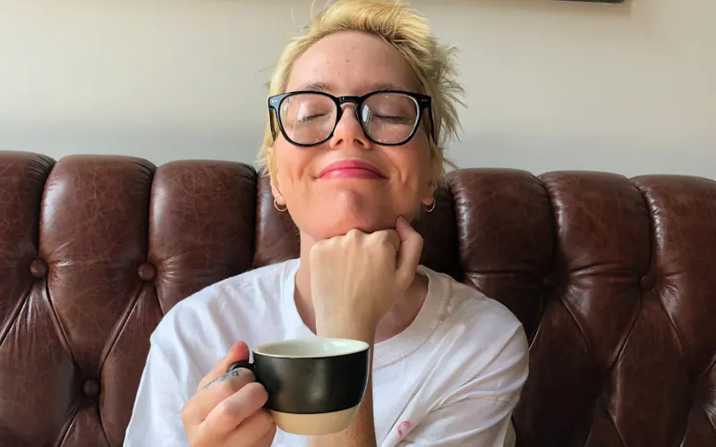 Amber is sitting on a leather couch, gently resting their head on their arm while holding a coffee cup.