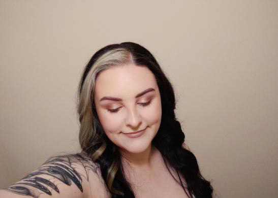 Beth is looking down and is smiling gently. Her hair is long and curled. Beth has a tattoo sleeve. 