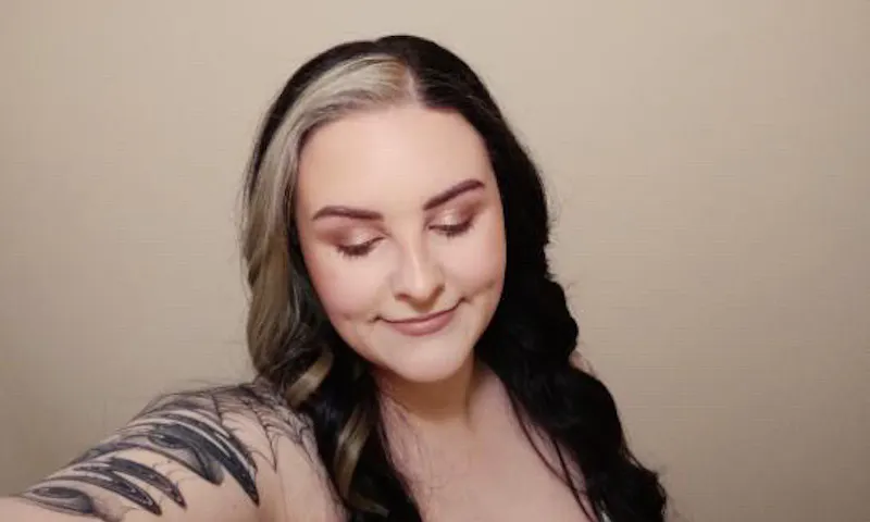 Beth is looking down and is smiling gently. Her hair is long and curled. Beth has a tattoo sleeve.