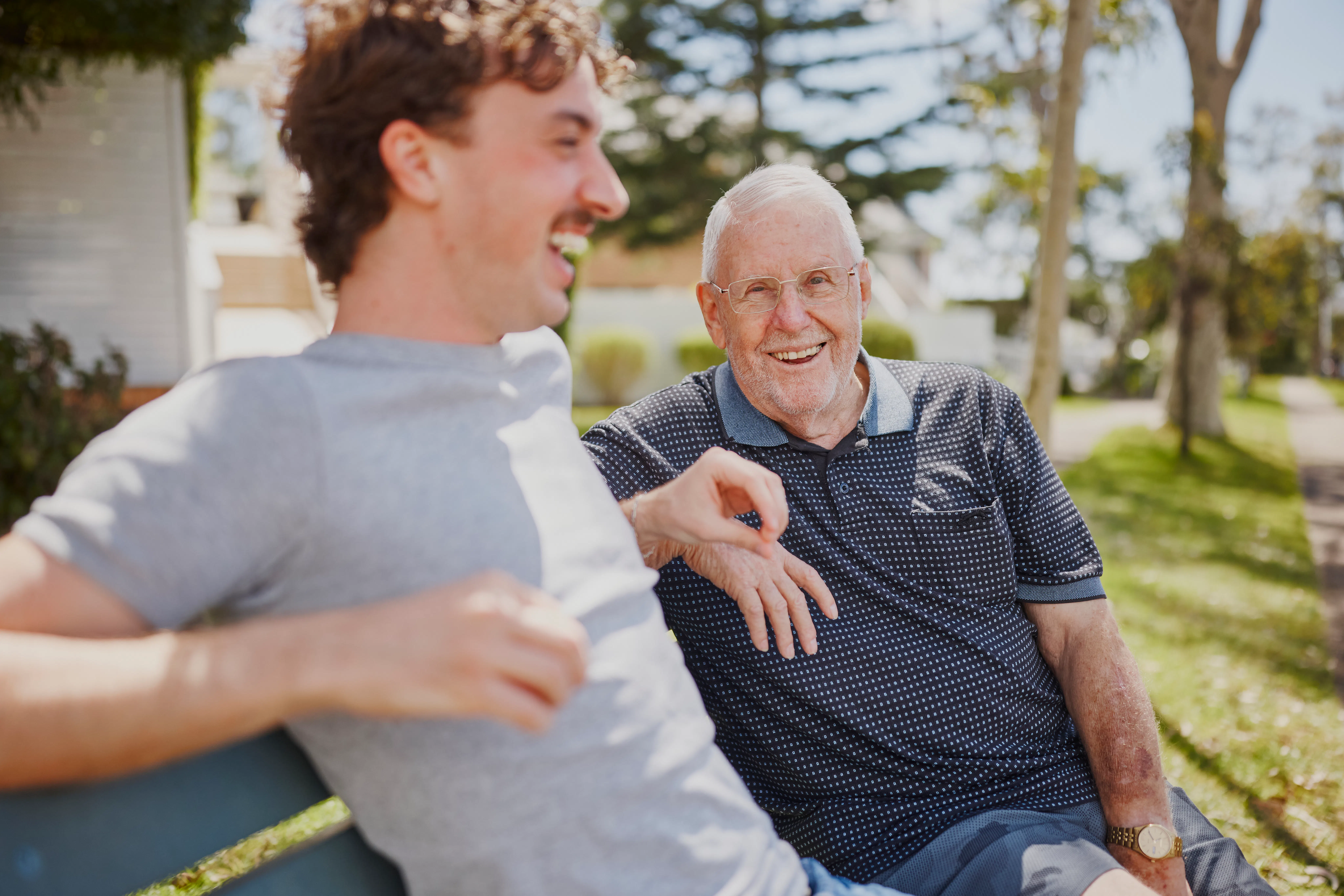 Frank and his support worker, Luka, are enjoying a sunny day together sitting on a park bench, laughing.