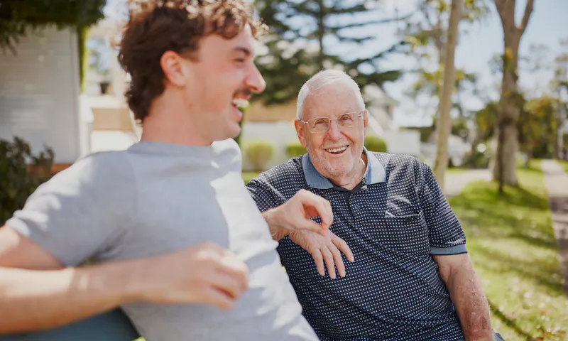 Frank and his support worker, Luka, are enjoying a sunny day together sitting on a park bench, laughing.