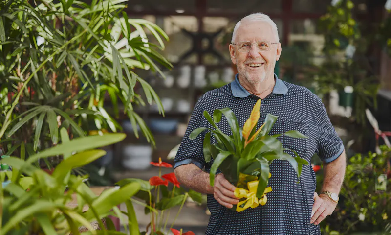 Frank is standing in a garden nursery, one hand resting on his hip while he holds a plant. He has short white hair and wears glasses.