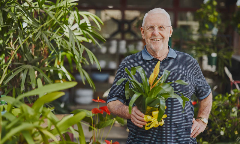 Frank is standing in a garden nursery, one hand resting on his hip while he holds a plant. He has short white hair and wears glasses.