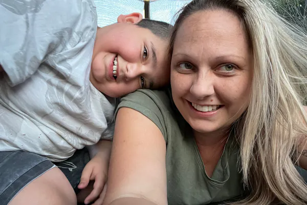 Amy is lying on a trampoline while her son, Luka, is resting his head on her shoulder. They're both smiling.