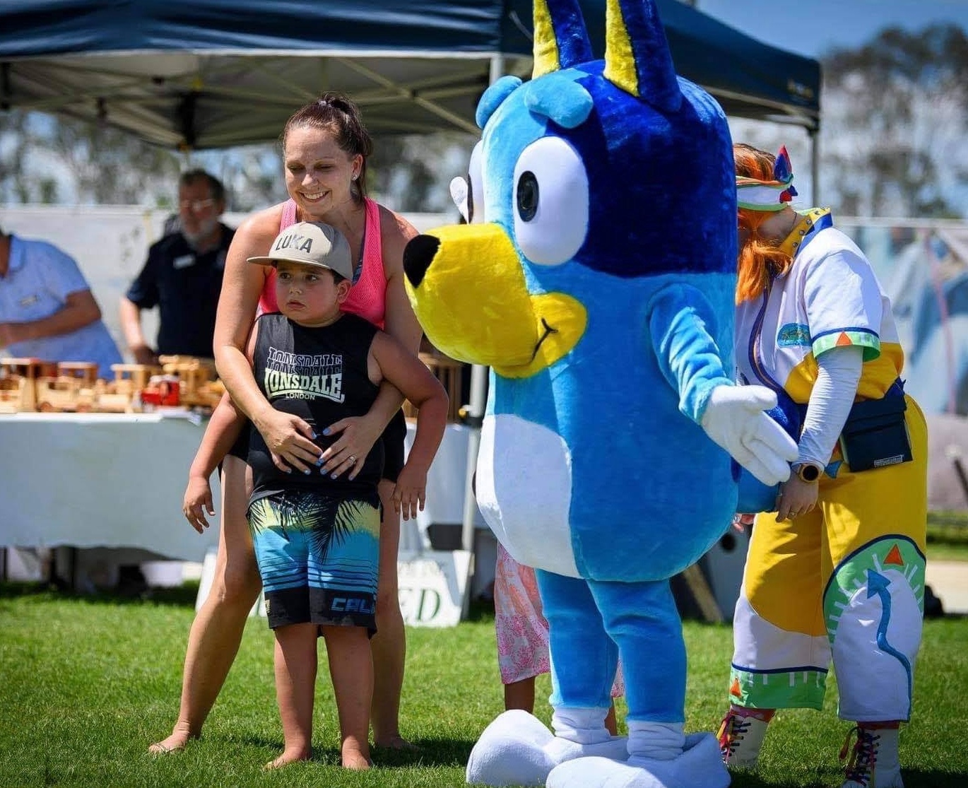 Amy's arms are wrapped around Luka. She's smiling and wearing a bright pink singlet. Luka wears a cap, singlet and board shorts. He has a cautious look on his face. A person wearing a large Bluey costume is posing next to Amy and Luka. 