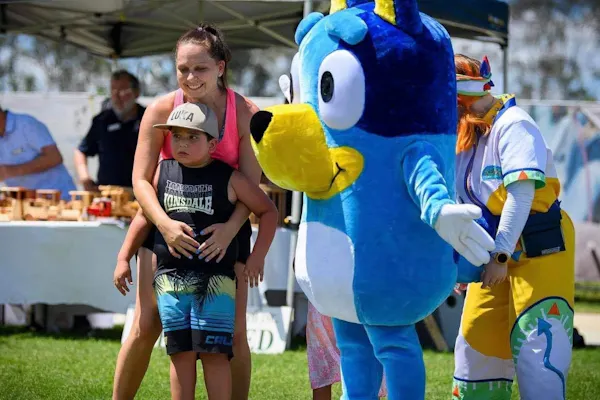Amy's arms are wrapped around Luka. She's smiling and wearing a bright pink singlet. Luka wears a cap, singlet and board shorts. He has a cautious look on his face. A person wearing a large Bluey costume is posing next to Amy and Luka.