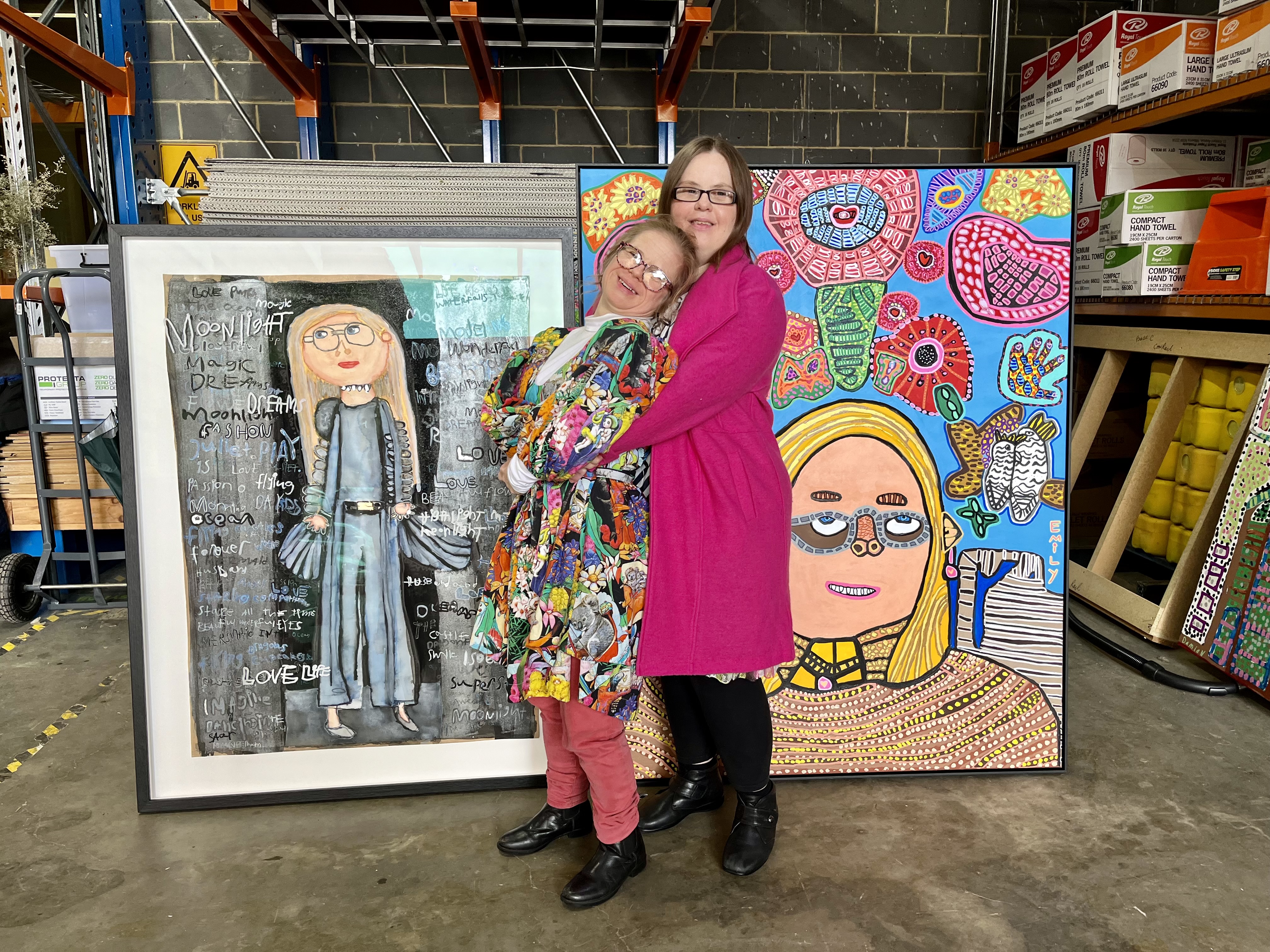 Studio A artists, Meagan Pelham and Emily Crockford, embrace in front of their Archibald Prize portraits. They're in a warehouse and both wear colourful outfits.