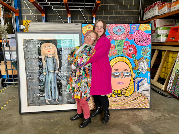 Studio A artists, Meagan Pelham and Emily Crockford, embrace in front of their Archibald Prize portraits. They're in a warehouse and both wear colourful outfits.