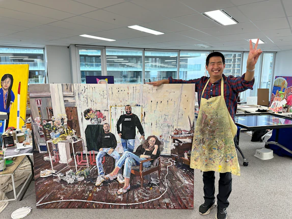 Studio A artist, Daniel Kim, proudly shows his Archibald Prize finalist painting. The painting is of three people in denim and black shirts in an art workshop. Daniel smiles while doing a 'peace sign', he's wearing an apron covered in paint.