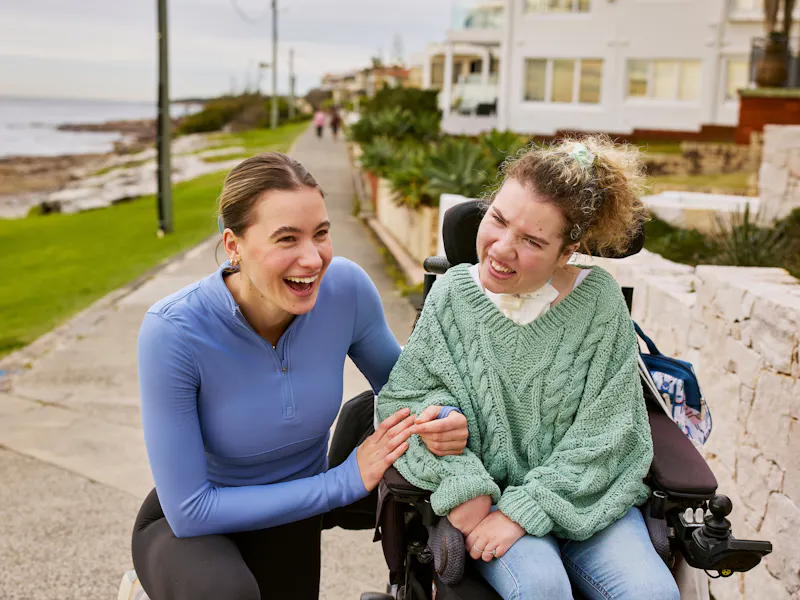 Hireup support worker Jane is kneeling beside Kiera's wheelchair. They smile with their arms linked. Kiera wears a sage knit jumper, while Jane wears a lilac activewear top. They are on a paved beach walk. Beachfront houses and a grey-looking sea appear in the background.