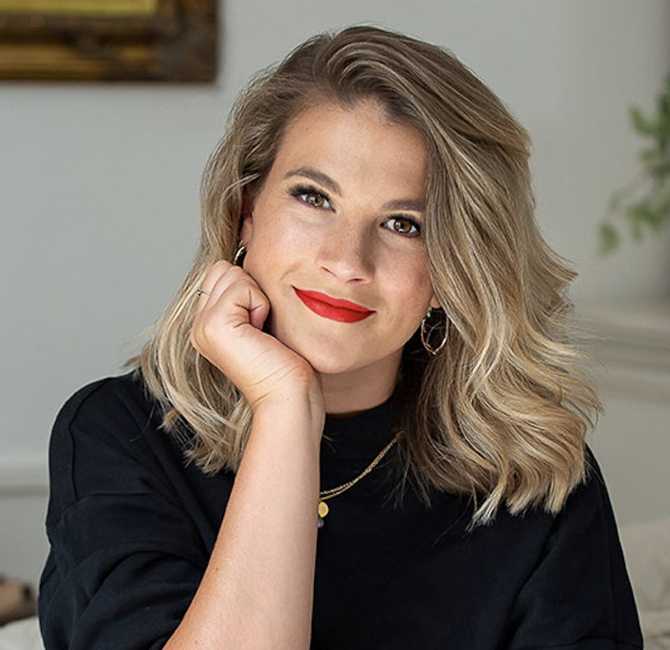 Grace props her chin in the palm of her right hand. She has shoulder-length blonde hair, wears a gold necklace and earrings, and wears a black top. Grace smiles warmly and wears red lipstick.