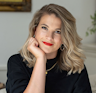 Grace props her chin in the palm of her right hand. She has shoulder-length blonde hair, wears a gold necklace and earrings, and wears a black top. Grace smiles warmly and wears red lipstick.