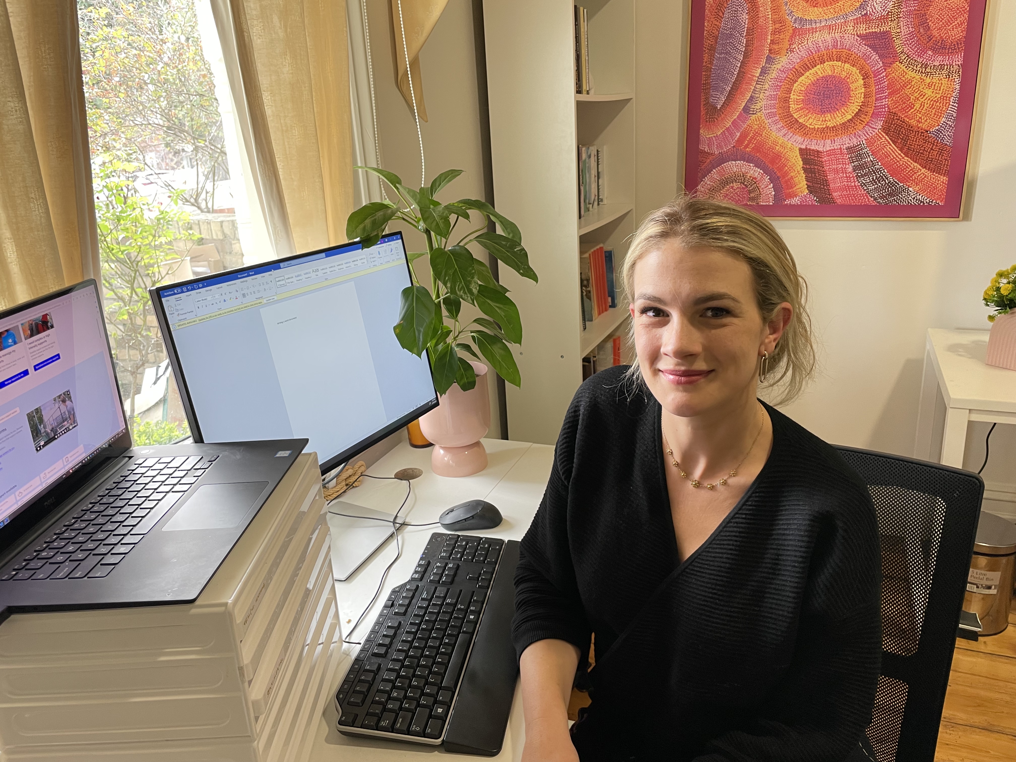 Grace is sitting at the desk of her home office. A desktop computer and laptop are on the desk. Sunlight peers in from the window and Grace smiles gently, she wears a black top and gold jewellery. Her office's bookshelf is full and a painting hangs on the wall. 