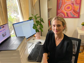 Grace is sitting at the desk of her home office. A desktop computer and laptop are on the desk. Sunlight peers in from the window and Grace smiles gently, she wears a black top and gold jewellery. Her office's bookshelf is full and a painting hangs on the wall.