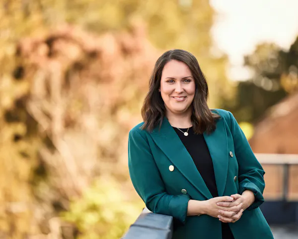Laura is outside on a balcony. She has dark, shoulder length hair and is wearing a green blazer. She's gently clasping her hands and is smiling.