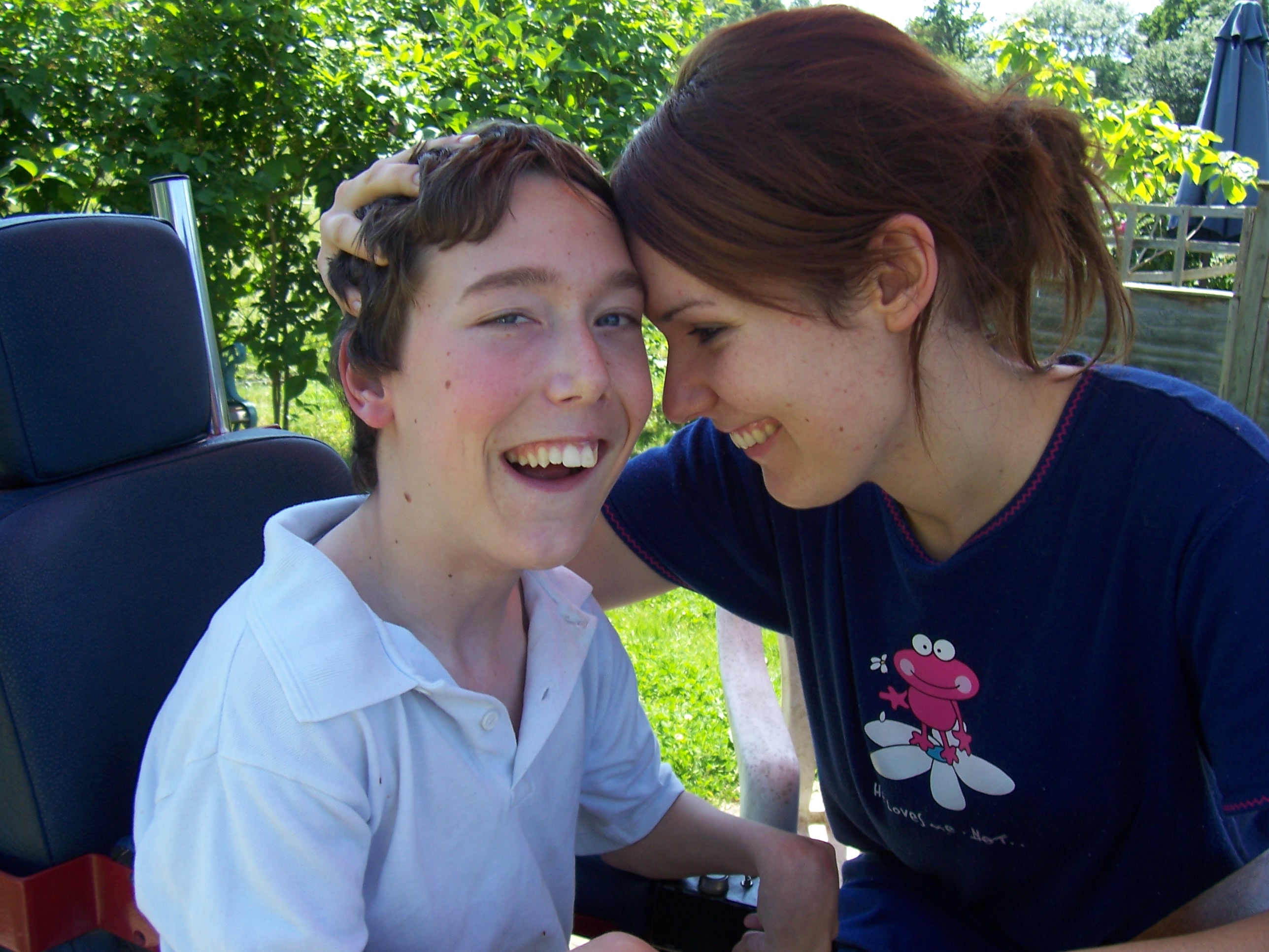 Laura and her younger brother Shane are in a garden. Laura is wearing a navy top with her hair back, smiling. She is gently bumping her forehead against Shane's, who uses a wheelchair. Shane is wearing a white polo top and is smiling. 