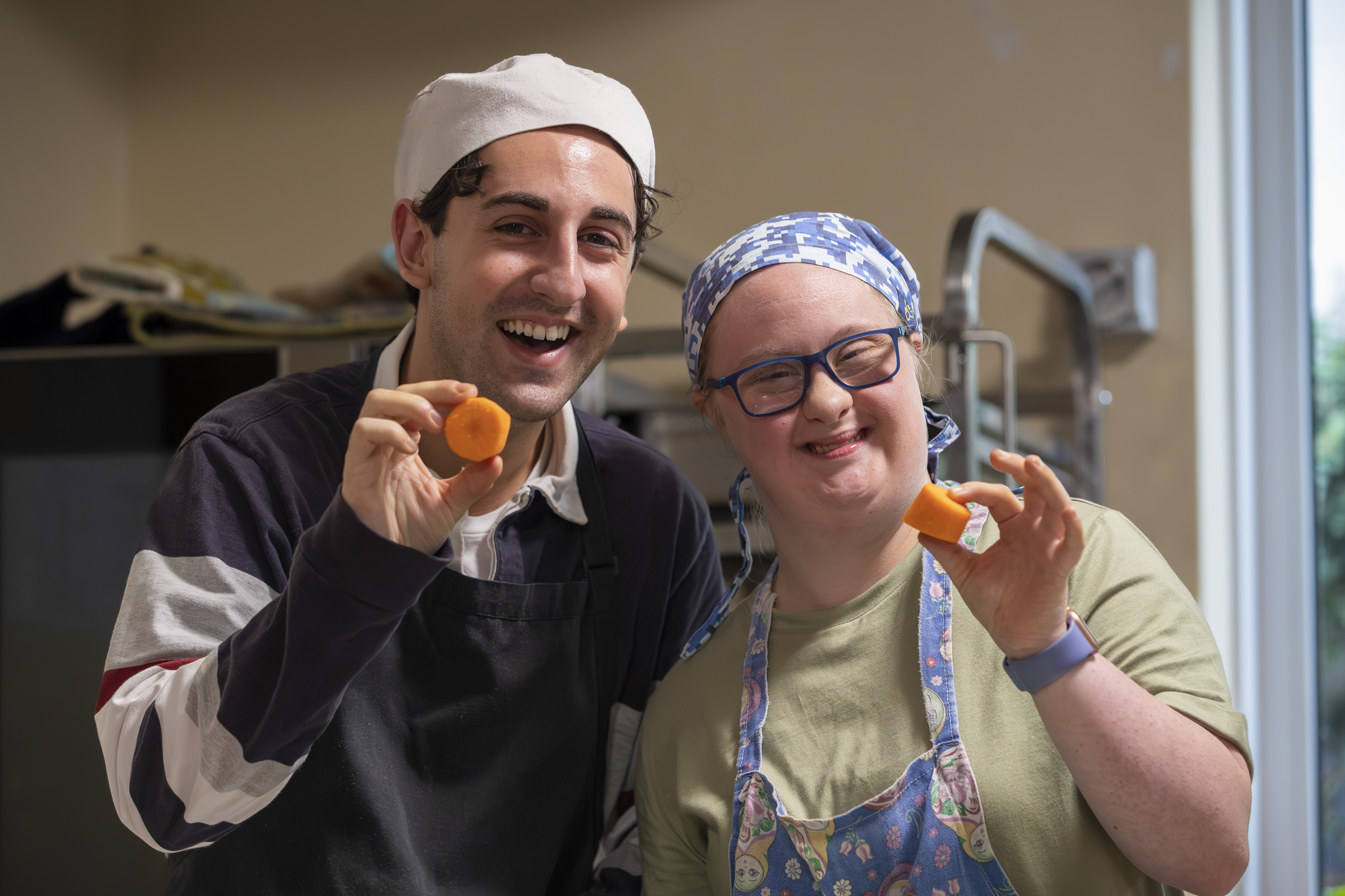 Hireup client Amy and her support worker Zak holding up chopped carrots in an industrial kitchen. They're both wearing hair nets and smiling at the camera.