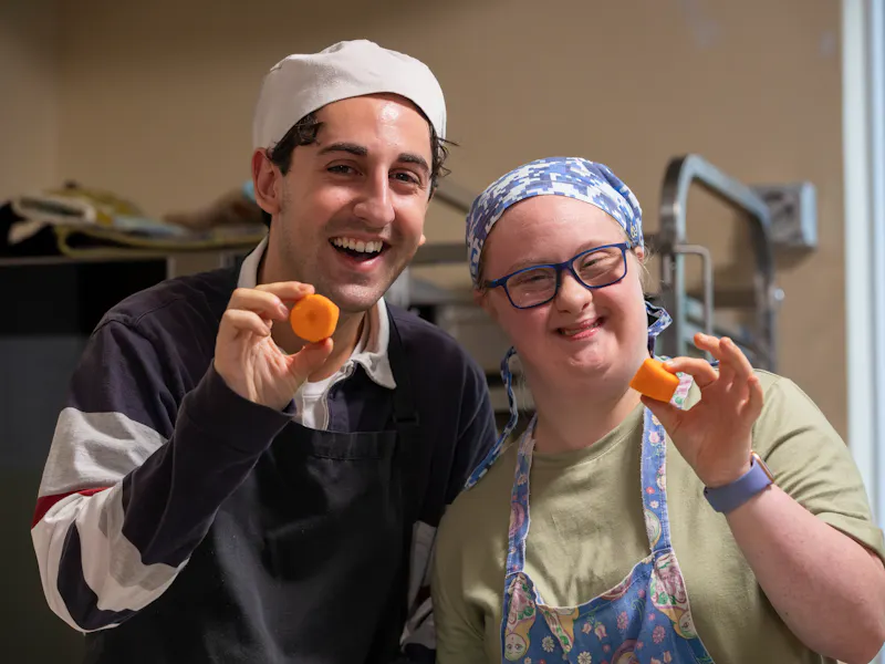 Hireup client Amy and her support worker Zak holding up chopped carrots in an industrial kitchen. They're both wearing hair nets and smiling at the camera.