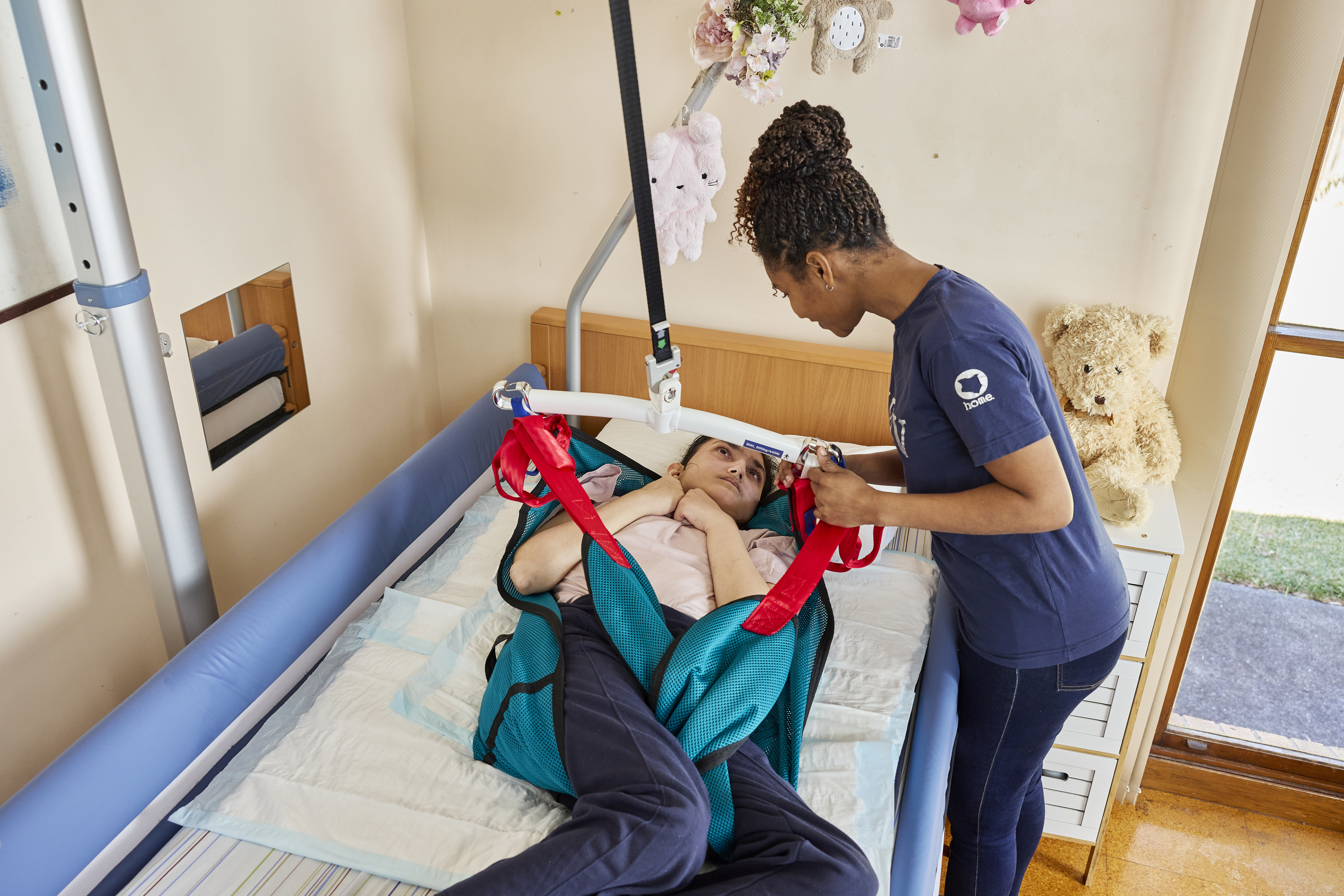Rifa is resting on a bed with a sling around her. She looks at her support worker, Mariam, and smiles. Mariam is carefully holding the hoist with one hand while gently guiding Rifa's head with the other.