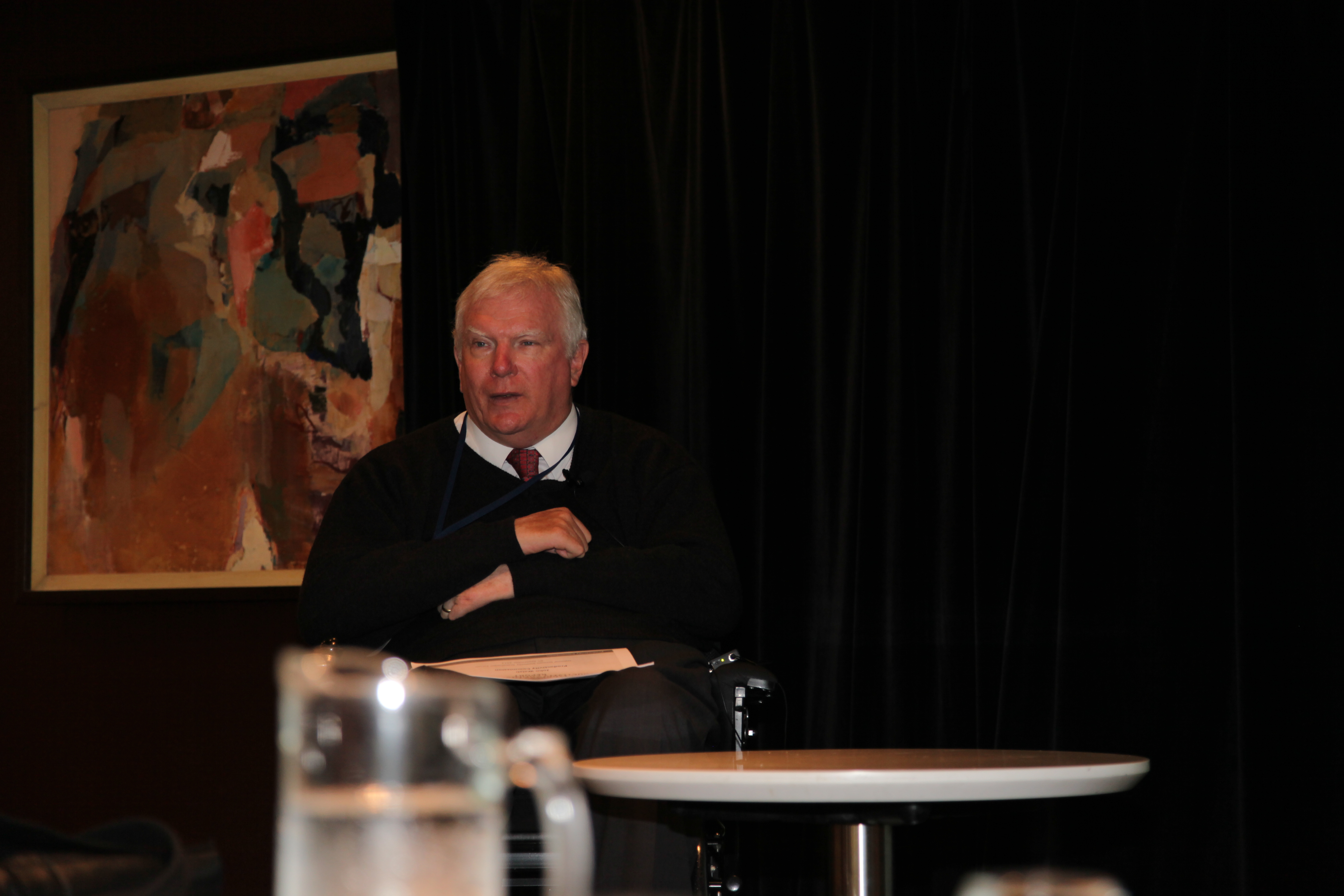 John Walsh has white hair and is wearing a black jumper. There is an abstract painting on the wall behind him. John uses a wheelchair.