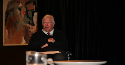 John Walsh has white hair and is wearing a black jumper. There is an abstract painting on the wall behind him. John uses a wheelchair.