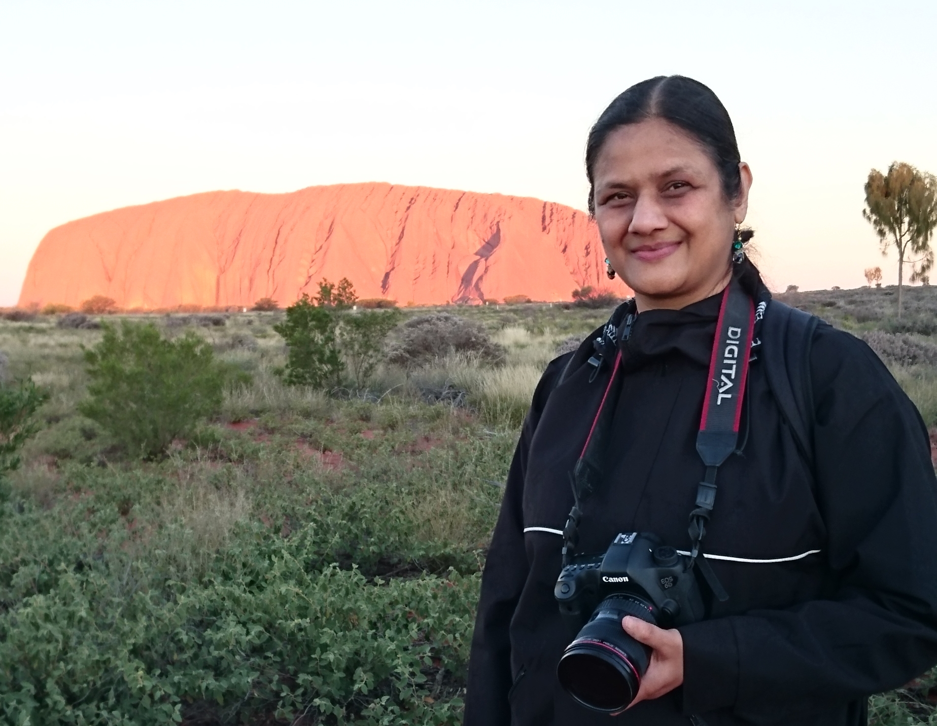 Journalist Neena Bhandari stands in front of Uluru holding a camera. 