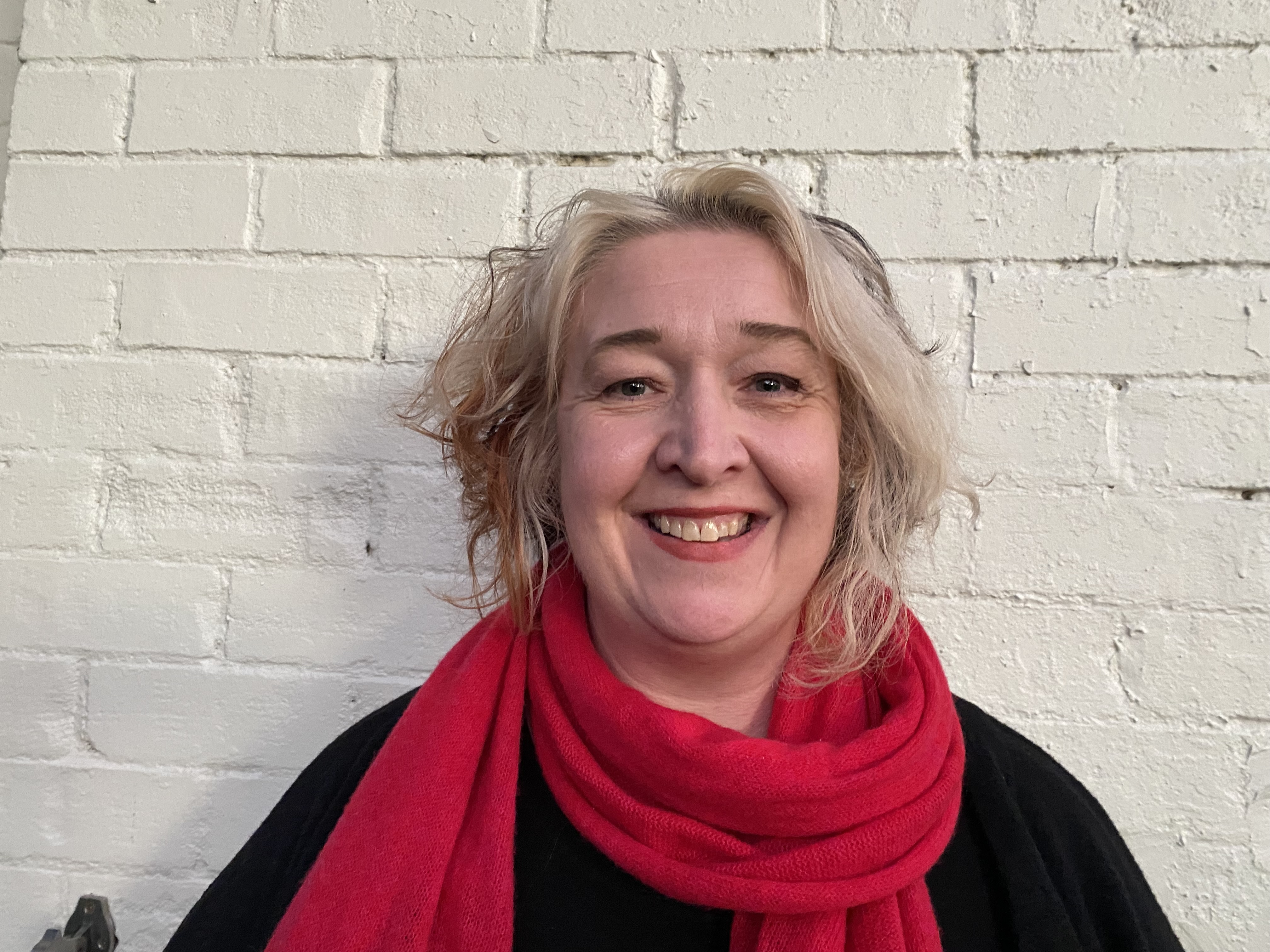 Kirsty is wearing a red scarf and is smiling widely. She's standing in front of a brick wall. 