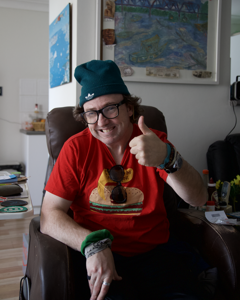 Goran is sitting in a brown armchair, smiling and giving a thumbs up. He is wearing a green beanie, reading glasses and a red t-shirt. 