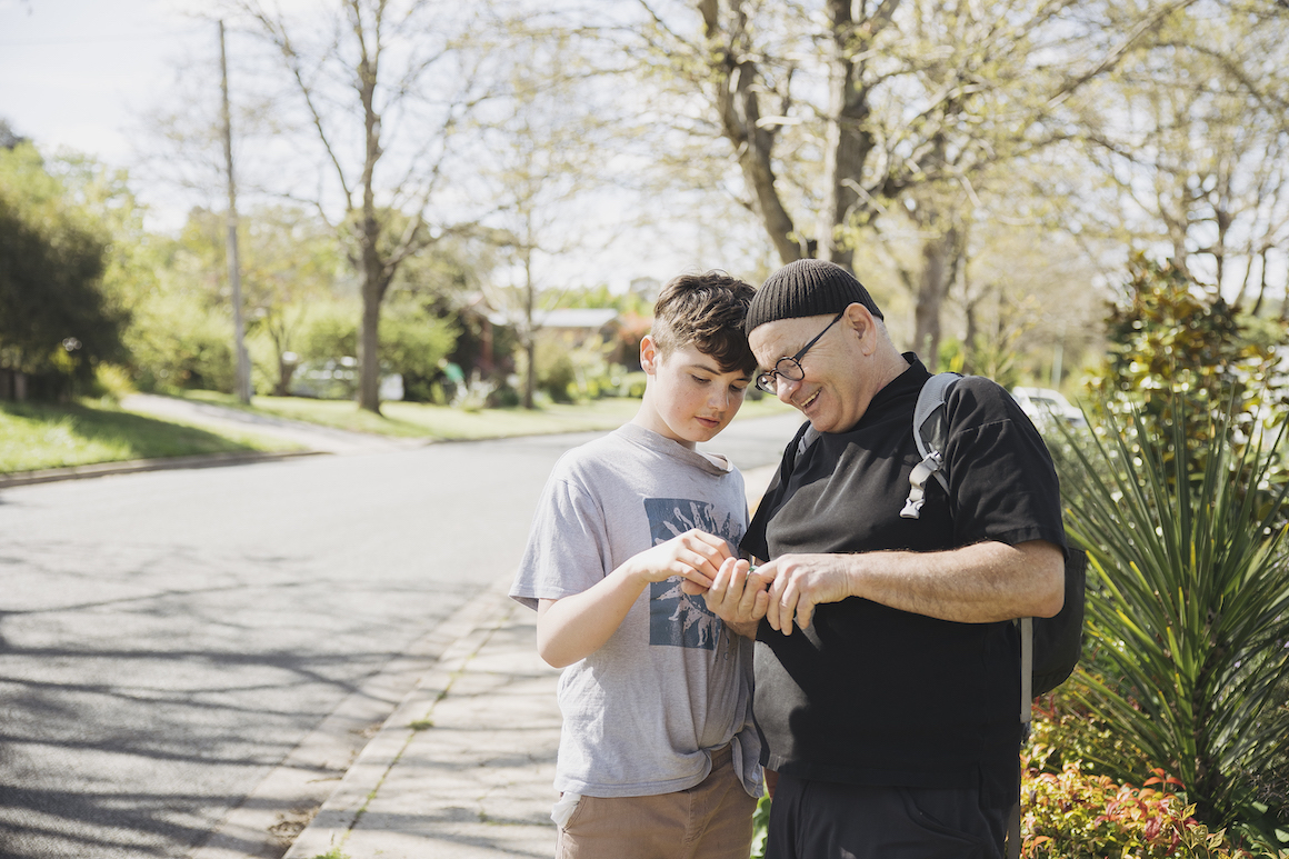Hireup client Zeph and his support worker Marcin are standing on a street pavement. Marcin is holding something in his hand, and both are smiling and inspecting it.