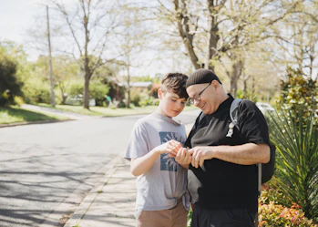 Hireup client Zeph and his support worker Marcin are standing on a street pavement. Marcin is holding something in his hand, and both are smiling and inspecting it.