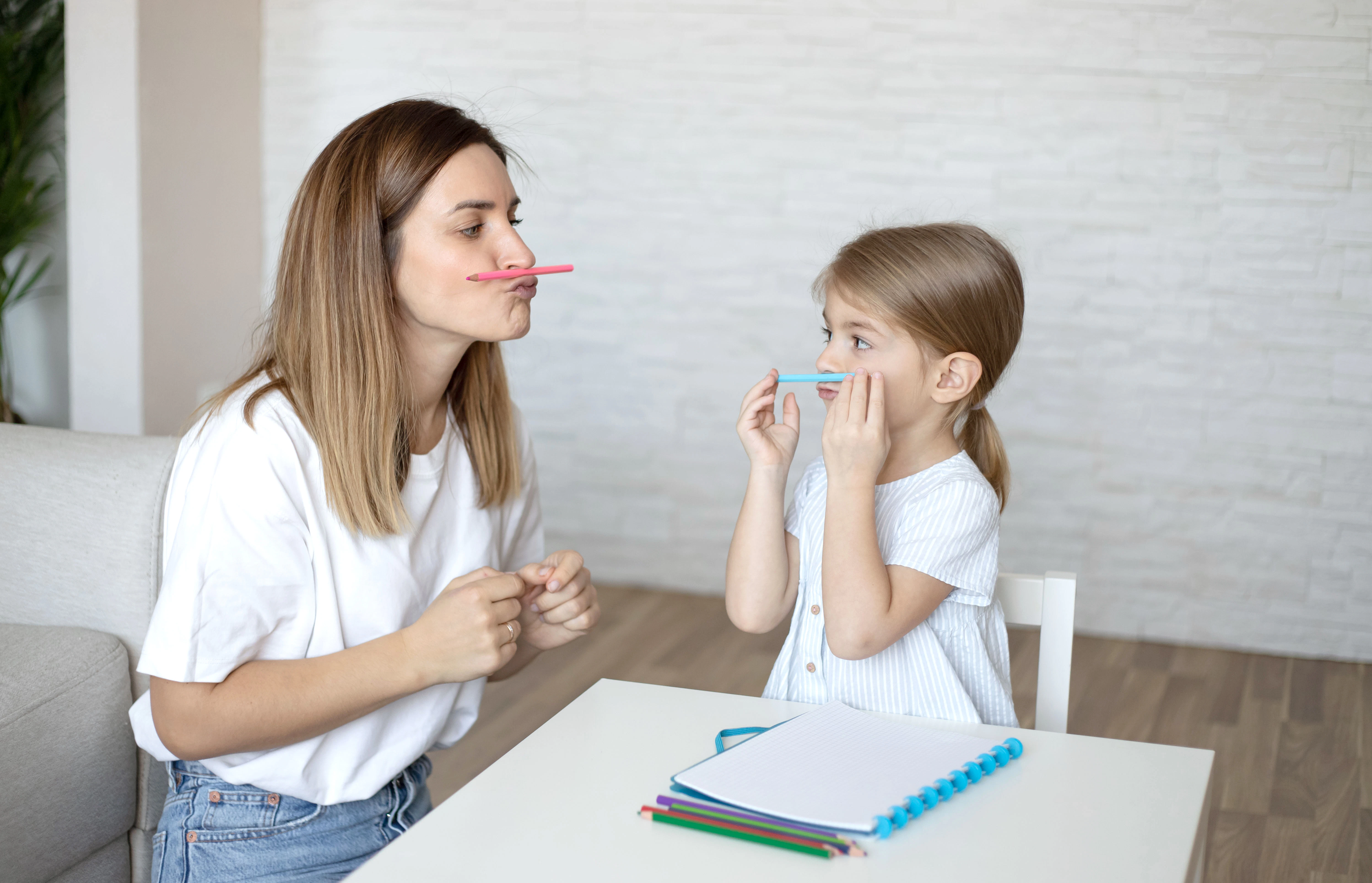 A female speech pathologist works with her client, a young girl. They are holding coloured pencils above their lips and looking at each other.