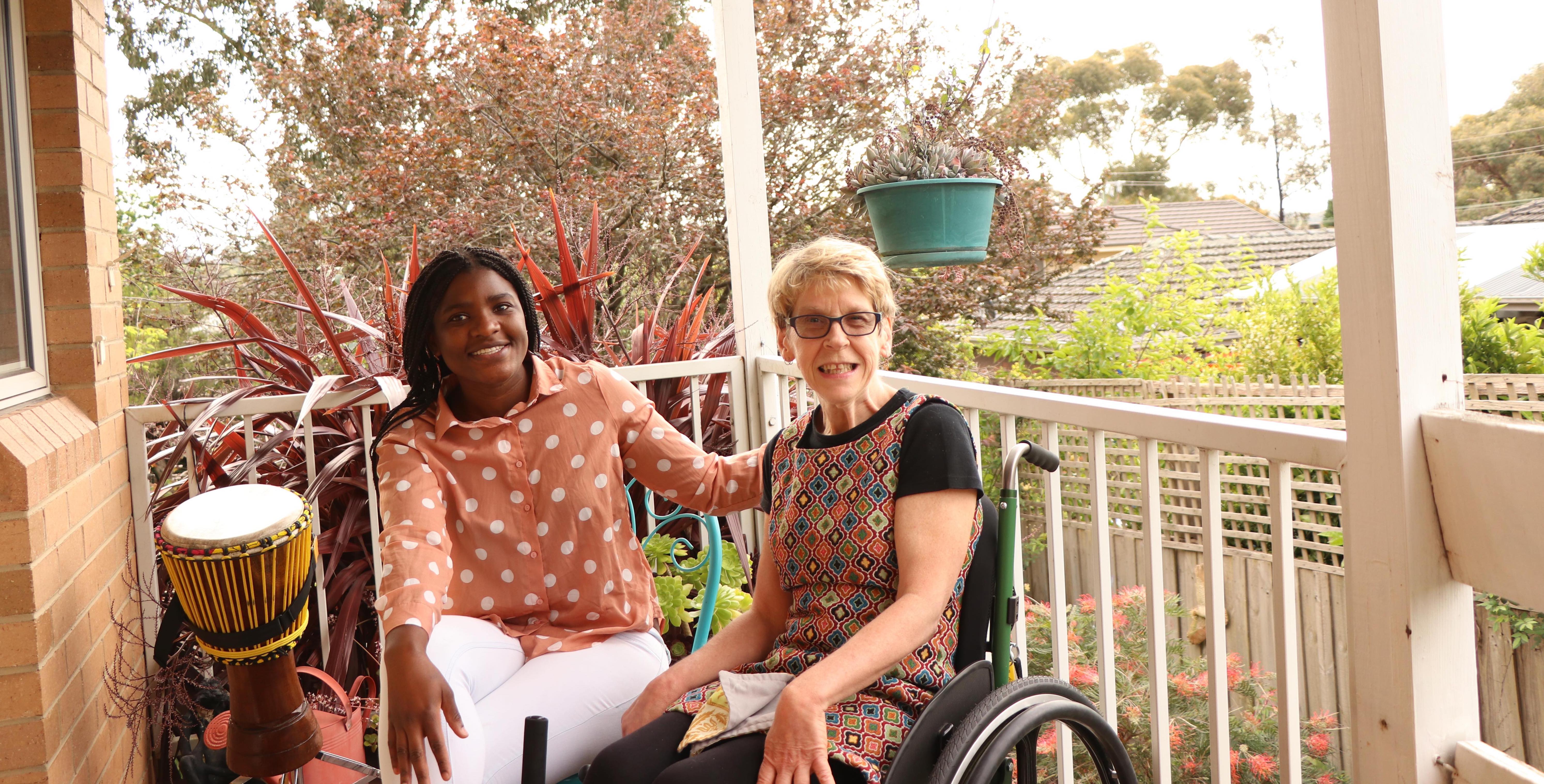 Idai and Melinda are on a veranda. They are both smiling and facing the camera. Idai has black hair that is styled in Micro-braids. She has her arm on Melinda's shoulder and there is a djembe (drum) beside Idai. Melinda has blonde hair and wears a patterned dress. Melinda is a wheelchair user. In the background, the garden is full of plants and trees.
