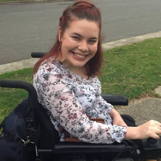 Nicole has short, red hair and is smiling. She's on a street path and is side on to the camera. Nicole is a wheelchair user. 
