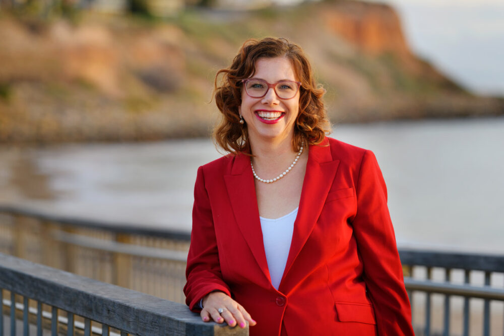 Labor Minister, Amanda Rishworth, is outside on a walkway near the water. She's smiling and wears a red blazer. 