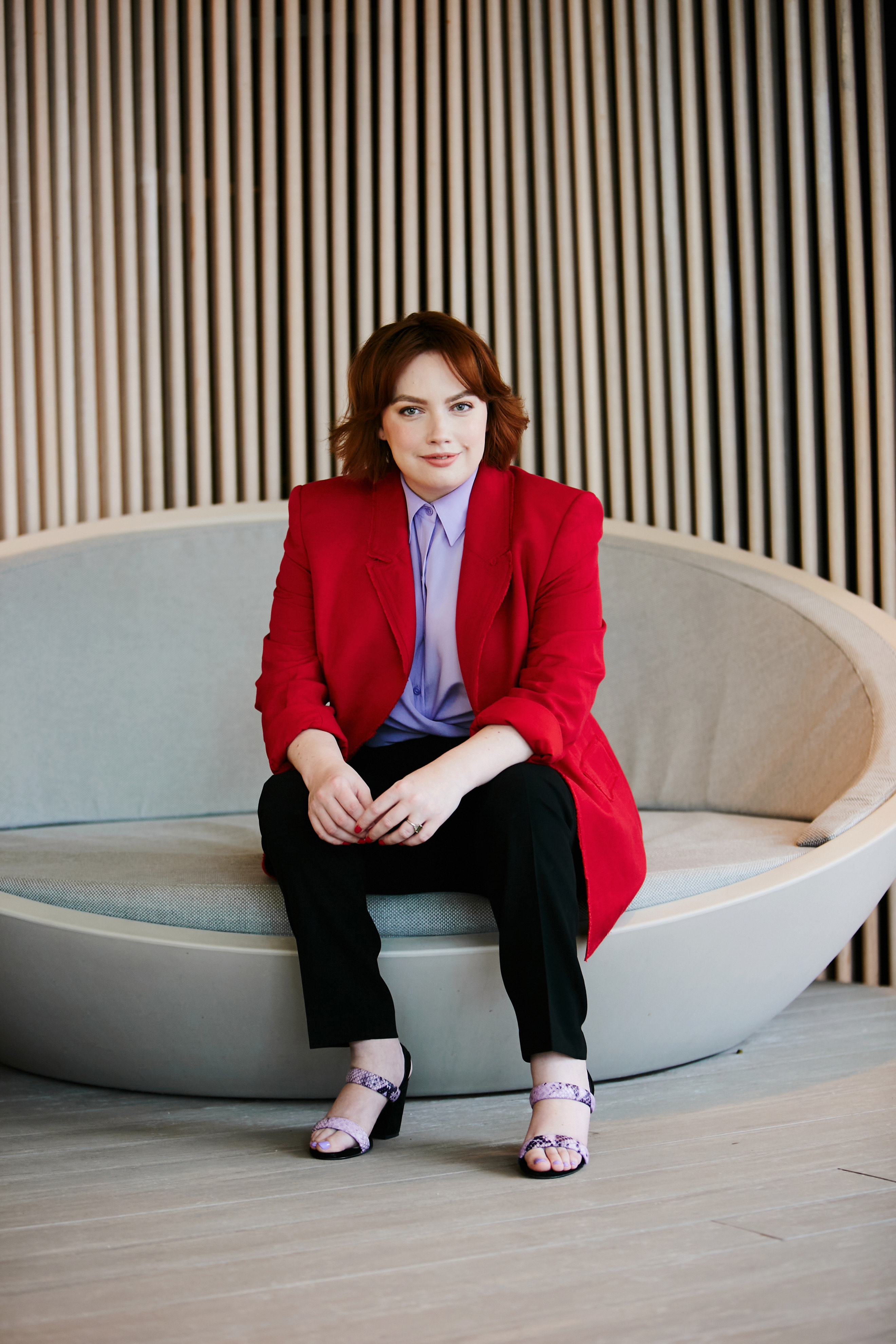 Disability rights advocate Elly Desmarchelier has shoulder-length red hair and wears a red blazer and purple shirt. She smiles at the camera and poses in a relaxed seated position.