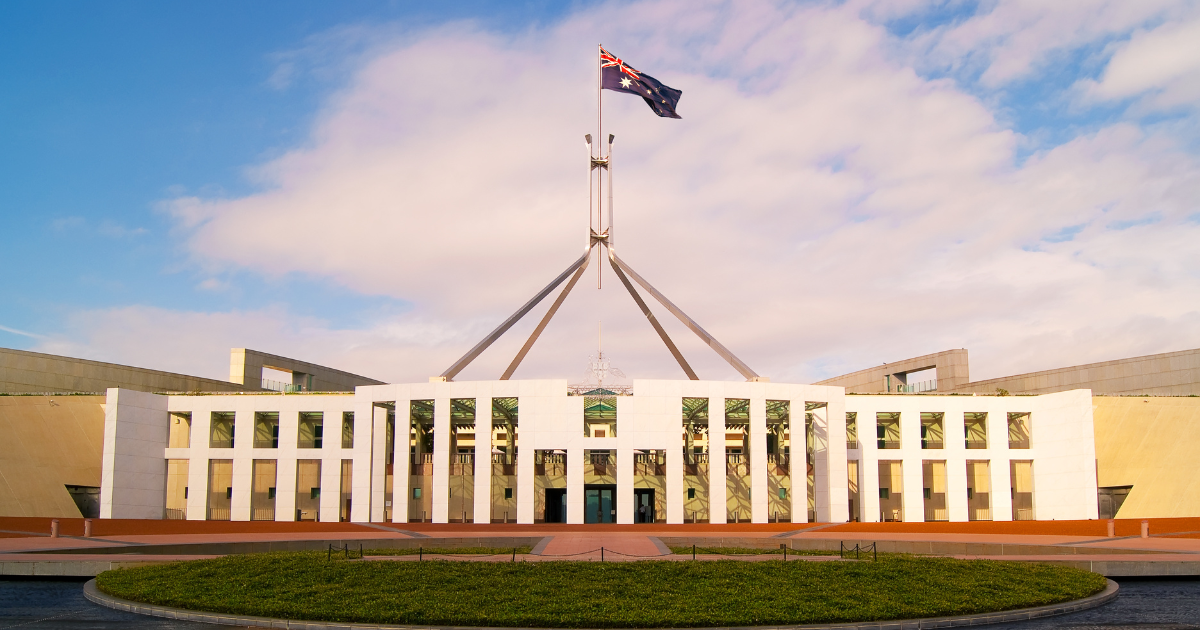 Australia's Parliament House is a wide white building with a wireframe of the country's coat of arms is nestled on top of the building. Australia's national flag also waves above the building.