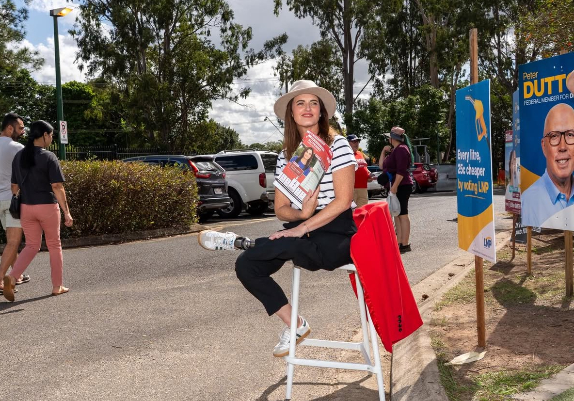 Labor Party's Ali France is sitting on a stool handing out flyers in the sunshine, smiling. Ali has a prosthetic leg.