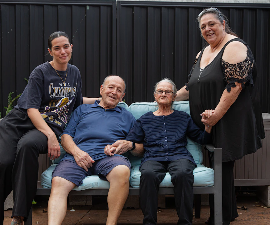 A Hireup aged care client, Victoria, is surrounded by her family. They are sitting outdoors.