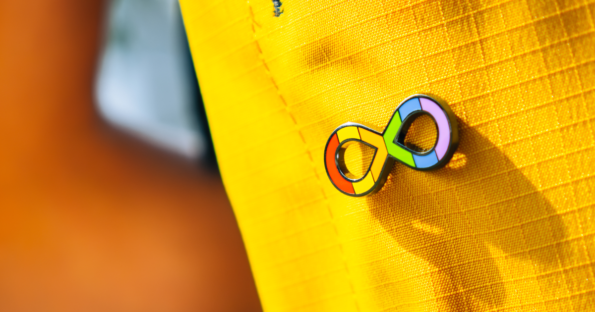 A pin is attached to a yellow item of clothing. The pin is a rainbow infinity symbol, which is widely recognised as the symbol for Autistic people.