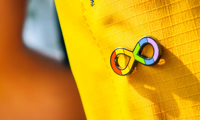 A pin is attached to a yellow item of clothing. The pin is a rainbow infinity symbol, which is widely recognised as the symbol for Autistic people.