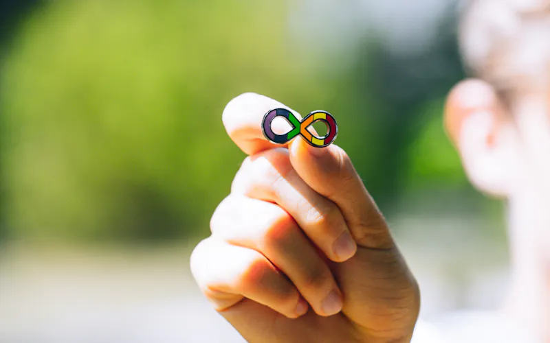 A person is holding onto an 'rainbow infinity symbol' pin. The symbol represents the spectrum of strengths, challenges and unique qualities of each Autistic person.