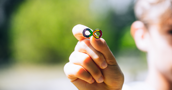 A person is holding onto an 'rainbow infinity symbol' pin. The symbol represents the spectrum of strengths, challenges and unique qualities of each Autistic person.