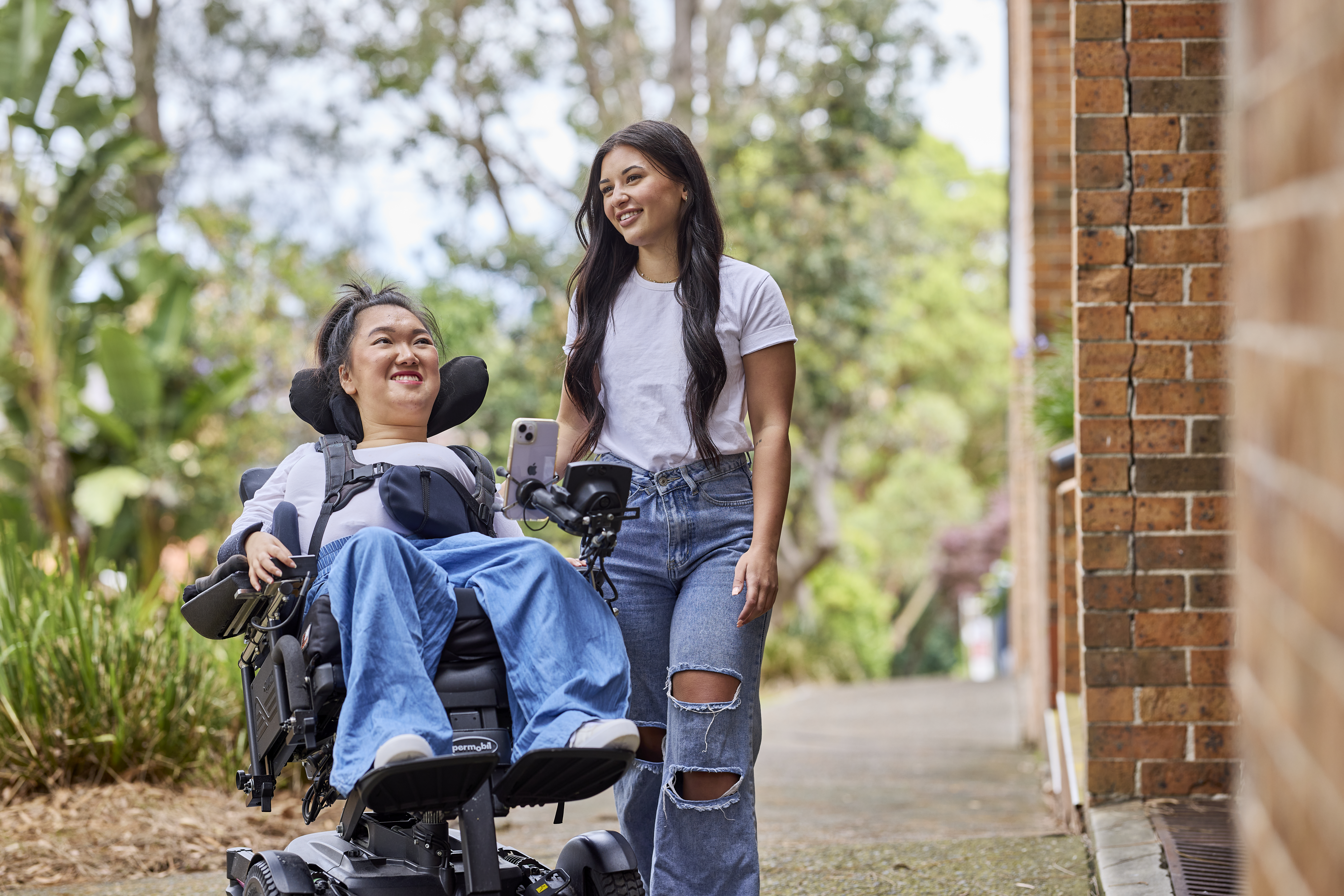 Melanie Tran and her support worker, Allyah, are strolling on a city path. Both are dressed in white t-shirts and denim jeans. Melanie is a wheelchair user.