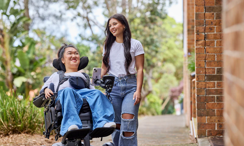 Melanie Tran and her support worker, Allyah, are strolling on a city path. Both are dressed in white t-shirts and denim jeans. Melanie is a wheelchair user.