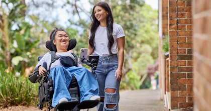 Melanie Tran and her support worker, Allyah, are strolling on a city path. Both are dressed in white t-shirts and denim jeans. Melanie is a wheelchair user.